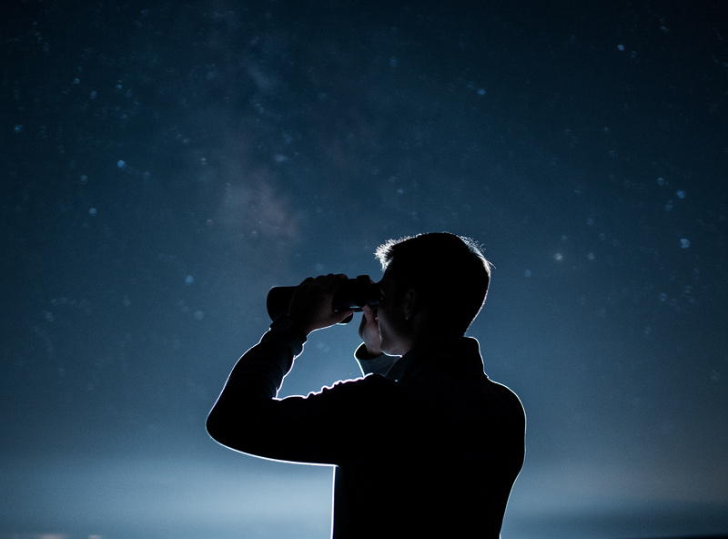 A person holding image-stabilized binoculars up to the night sky, hands steady, stars overhead