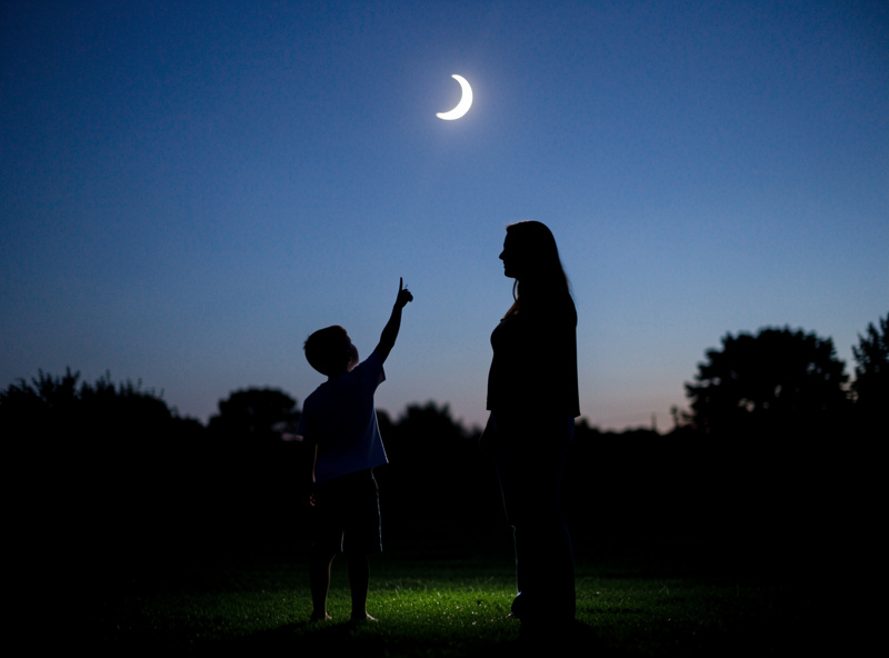 A child pointing up toward a bright crescent Moon at dusk with a parent beside them