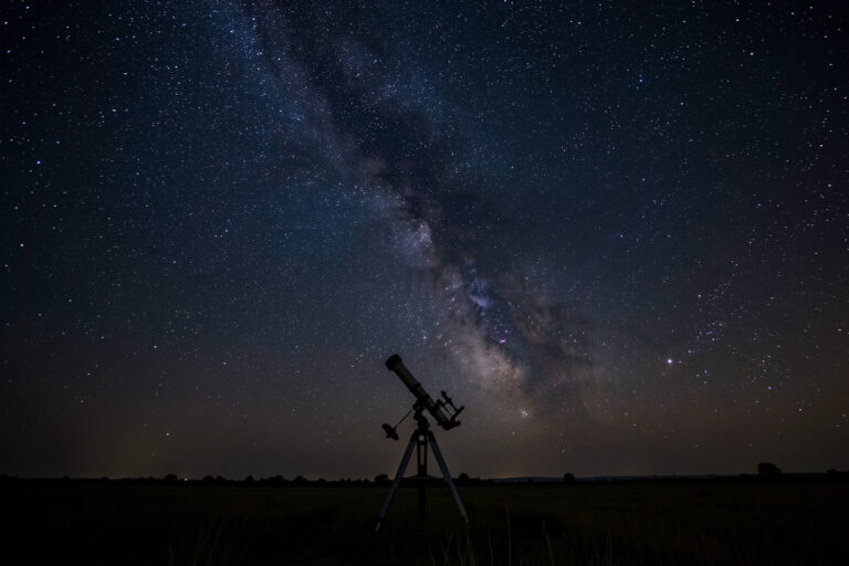 Dark starry night sky and faint Milky Way over a silhouetted telescope in a rural field