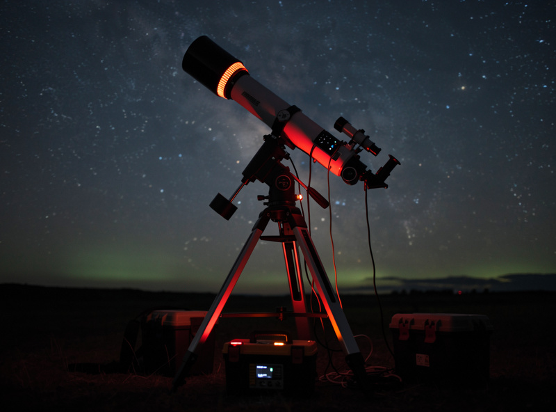 Astrophotography telescope rig on equatorial mount in dark field under stars, dew heater strip visible on corrector plate, battery pack at tripod base